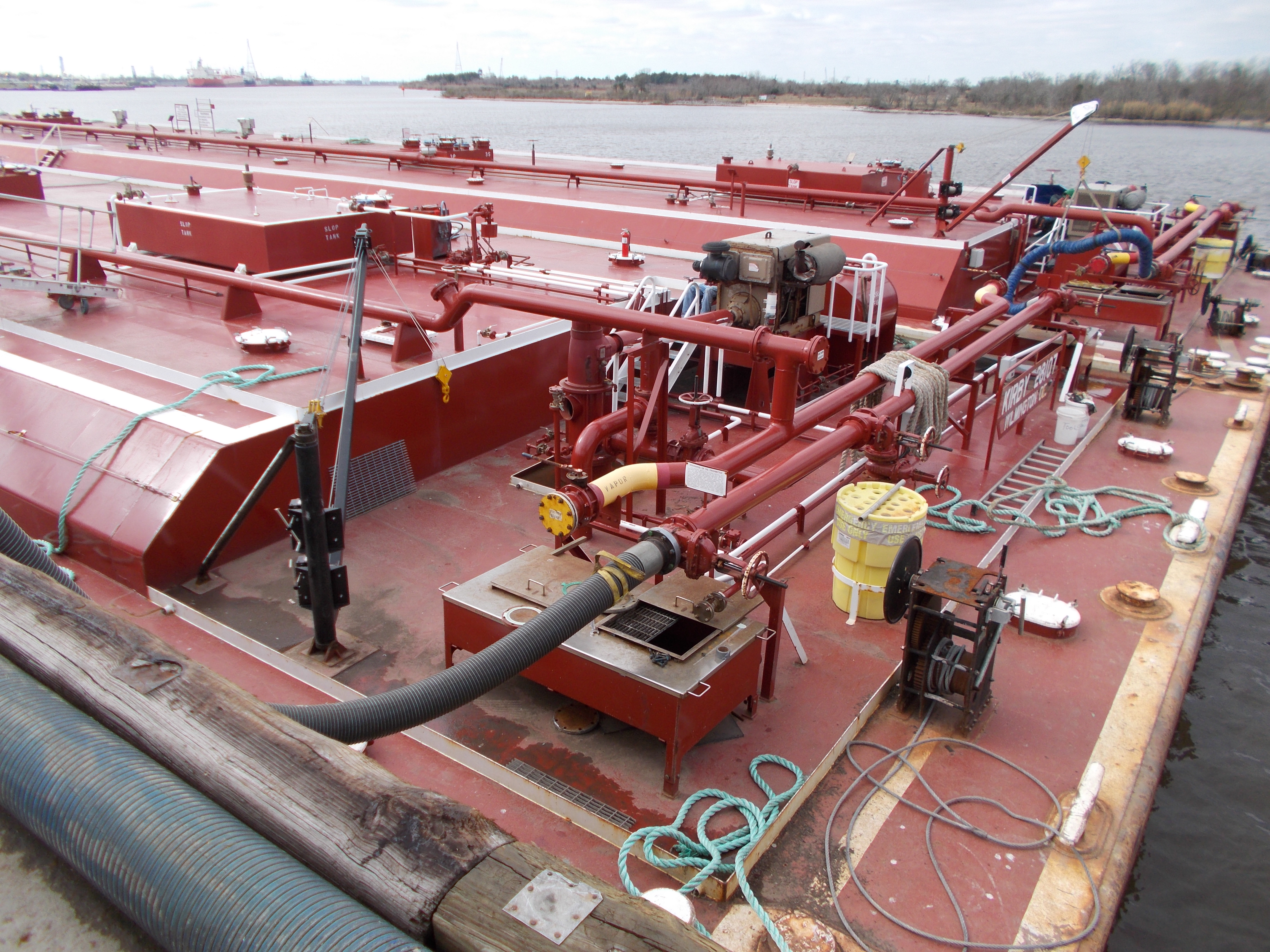 Deck of a red tank barge during transfer — hoses, winches, mooring ropes, Ohio River background